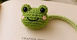Close-up of a green amigurumi frog head bookmark topper with black eyes and pink cheeks